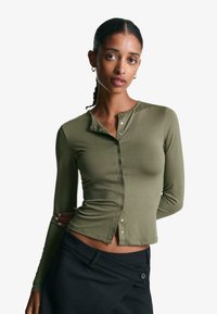 Maglietta a maniche lunghe verde oliva con dettagli in bottone, caratterizzata da un design aderente e una superficie liscia. Abbinata a pantaloni neri.