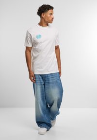 Hvit bomull t-skjorte med et turkist grafisk motiv på brystet. Modellen har på seg løstsittende, lyseblå denimjeans med en falmet effekt.