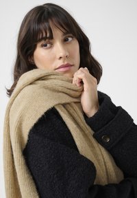 Un manteau noir texturé avec un large col et des poignets boutonnés, drapé d'une douce écharpe beige, mettant en valeur des matériaux chauds et superposés.