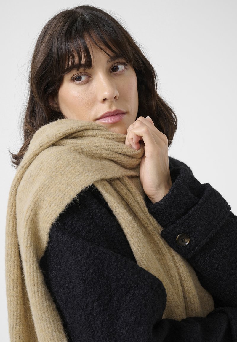 Un manteau noir texturé avec un large col et des poignets boutonnés, drapé d'une douce écharpe beige, mettant en valeur des matériaux chauds et superposés.