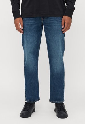 Person iført blå lige ben jeans kombineret med sorte sneakers og en sort langærmet skjorte, stående mod en ensfarvet baggrund.