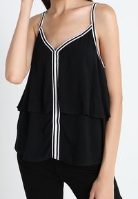 Top senza maniche nero con un design a strati, caratterizzato da un scollo a V e rifiniture bianche lungo le spalline e la cucitura centrale.