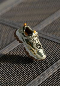 Zapatillas deportivas verdes y beige con suela texturizada, parte superior de malla, acentos amarillos y un logo prominente. Incluye cordones y una lengüeta trasera.