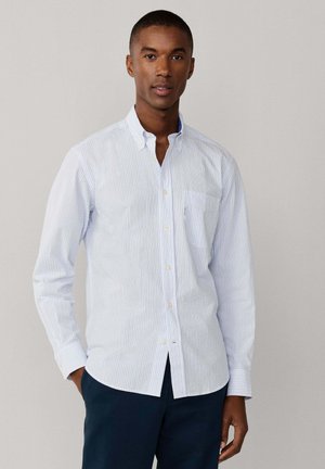 Homme portant une chemise à rayures bleu clair et blanches avec col et un pantalon bleu marine foncé, debout devant un fond uni.