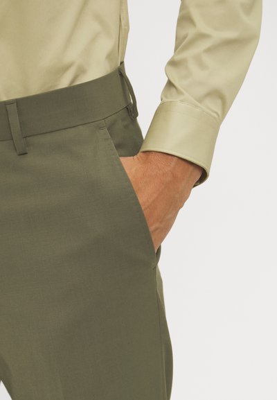 Schmal geschnittene olivgrüne Hose mit flachem Bund, Seitentaschen und einem glatten, leicht strukturierten Stoff. Kombiniert mit einem hellbeizigen Hemd.