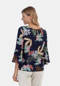 Blusa floreale navy con maniche a campana, motivi tropicali vivaci con foglie verdi e fiori rossi, e chiusura a bottone sul retro.