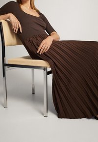 Braunes Kleid mit einem figurbetonten Oberteil und einem plissierten, fließenden Rock. Weiche Textur, lange Länge, kombiniert mit einem einfachen Stuhl mit hellholzfarbenem Sitz und Metallbeinen.