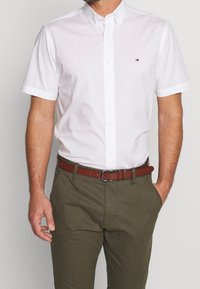 Uomo che indossa una camicia bianca a maniche corte, infilata in pantaloni verde oliva con una cintura marrone, con una mano in tasca.