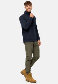 Mørkeblå zip-up sweatshirt med blød tekstur, parret med olivengrønne tapered bukser og brune snørebåndsstøvler med robuste såler.