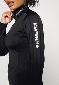 Schwarzes Sportoberteil mit Reißverschluss und hohem Kragen. Mit kontrastierendem weißen Logo am Arm und strukturiertem Material, taillierte Passform.