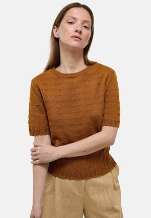 Femme à la peau claire et aux cheveux bruns raides porte un haut en maille à manches courtes couleur rouille et un pantalon beige rayé, debout devant un fond blanc.