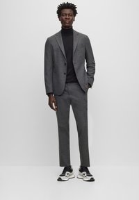 Costume gris texturé avec revers crantés, fermeture à un bouton, associé à un col roulé noir et des sneakers blanches avec des accents noirs.