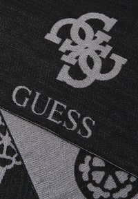 Juodas šalikas su išskirtiniu sidabriniu logotipu, kuriame yra persipynusios raidės ir žodis "GUESS" storomis raidėmis. tekstūruota medžiaga su geometriškais raštais.