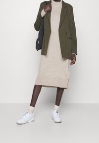 Blazer doppiopetto verde oliva, abito di maglia beige con orlo a coste, sneakers bianche e borsa a tracolla nera trapuntata.