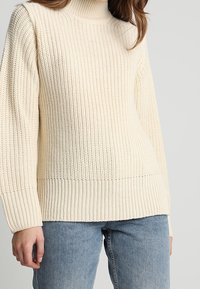 Cremfarbener, grob gestrickter Rollkragenpullover, getragen mit hellblauen Jeans, die den Oberkörper und eine Hand einer Person zeigen.