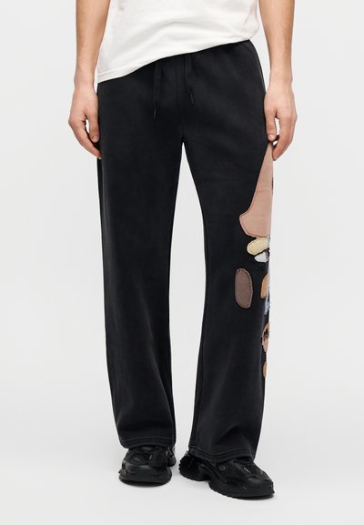 Μαύροι αθλητικοί παντελόνες με υφή, διακοσμημένοι με πολύχρωμα υφασμάτινα μπαλώματα και ελαστική μέση με κορδόνι. Συνδυάζονται με μαύρα αθλητικά παπούτσια.