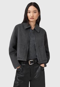 Veste grise en feutre avec fermeture éclair sur le devant, large col et deux poches avant. Portée sur un haut gris, associée à un pantalon en simili cuir noir et une ceinture.