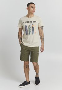 T-shirt beige avec un graphisme de Californie et des planches de surf colorées ; associé à un short cargo vert olive et des baskets noires.