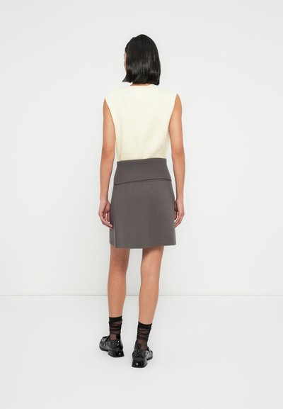 Femme debout, de dos, portant un haut blanc sans manches, une jupe grise longueur genoux avec une large ceinture, des chaussettes noires transparentes et des chaussures noires.