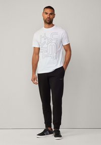 Witte T-shirt met grijs geometrisch patroon, gecombineerd met zwarte joggers met een logodetail. Zwarte sneakers met grijze accenten maken de outfit compleet.