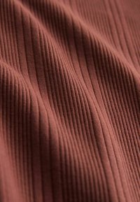 Rífľované tkanivo v teplej hnedej farbe, so zvislými prúžkami a jemnou textúrou. Materiál sa zdá byť elastický a hladký.