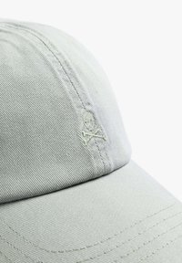 Lys grå baseballkasket lavet af struktureret stof, med et broderet dødningehoved og korslagte knogler detalje. Skyggen er buet med synlige sømme.