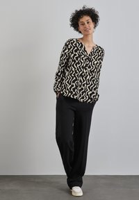 Schwarz-beige gemusterte Bluse mit langen Ärmeln, Knopfverschluss vorne, lockere Passform, kombiniert mit lockeren schwarzen Hosen und hellen Schuhen.