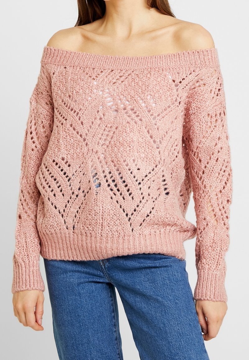 Jasnoróżowy, sweter robiony na drutach typu off-the-shoulder z teksturowanym otwartym wzorem, ściągaczami przy mankietach i u dołu. Nosi się go na niebieskie dżinsy.