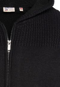 Czarna dzianinowa bluza z kapturem na zamek błyskawiczny, o prążkowanej teksturze i miękkim, puszystym wnętrzu. Posiada srebrny zamek i metkę na kołnierzu.