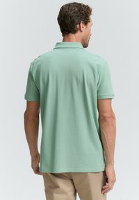 Mand iført lysegrøn kortærmet polo-shirt og beige bukser, stående med ryggen til foran en ensfarvet lys baggrund.