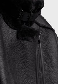 Black outerwear featuring textured fabric, a high collar, and adjustable leather strap. Includes a visible zip closure on one side.