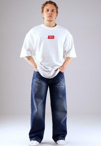 Junger Mann steht mit den Händen in den Taschen, trägt ein übergroßes weißes T-Shirt mit rotem Logo, lockere blaue Jeans und weiße Sneakers vor einem einfachen Hintergrund.