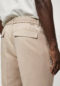 Beżowe spodnie z elastycznym pasem, z dwoma tylnymi kieszeniami i subtelną teksturą. Widoczna metka blisko pasa.