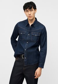 Άνδρας που φοράει σκούρο μπλε πουκάμισο από τζιν με κουμπιά τύπου pearl snap και μαύρο τζιν, στέκεται μπροστά σε απλό ανοιχτό φόντο.