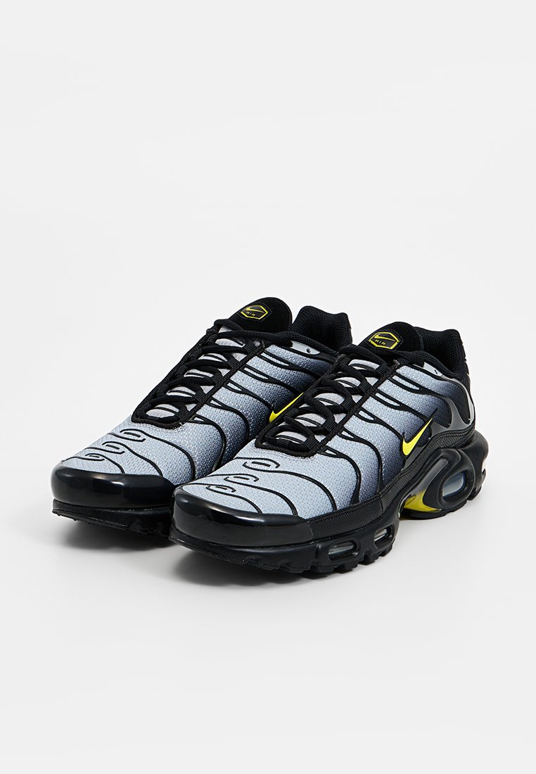 Baskets Nike avec une tige en maille grise, des accents noirs, un logo jaune, des lacets élastiques et une semelle noire arrondie avec un amorti d'air.