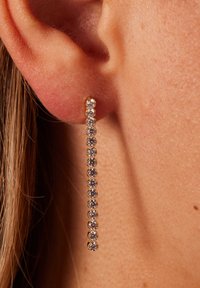 Goldene Tropfenohrringe mit einer geraden Linie aus klaren Kristallen, die eng beieinander gesetzt sind und unterhalb des Ohrläppchens vor einem strukturierten Hauthintergrund hängen.