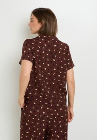 Chemise à manches courtes bordeaux avec un motif léopard comportant des taches beige et noires. Tissu lisse avec une coupe décontractée et un col.