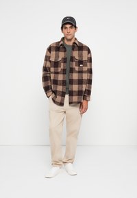 Man in een zwart en bruin geruit jasje, groen sweater, beige broek, witte sneakers en een donkere baseballpet, staand tegen een witte achtergrond.