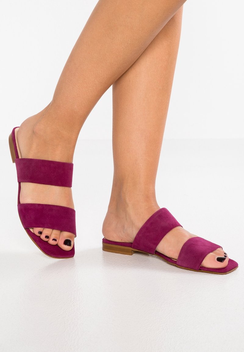 Nohy obuté v plochých, otevřených fialových semišových pantoflích s dvěma širokými pásky a černým lakem na nehty na bílém podlaze.