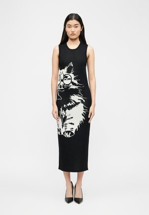 Femeie purtând o rochie midi neagră fără mâneci, cu o grafică mare a unei pisici albe cu ochelari, asortată cu pantofi negri cu vârf ascuțit.