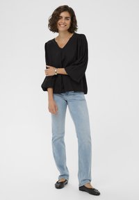 Blouse noire à col en V et manches longues avec des accents froncés, associée à un jean bleu clair et des ballerines noires. Modèle se tenant sur un fond uni.