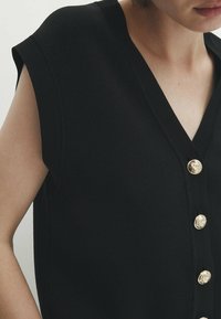 Mustaton hihaton toppi V-pääntiellä, jossa on kultaiset napit edessä, valmistettu sileästä materiaalista. Minimalistinen muotoilu ja rakenne.