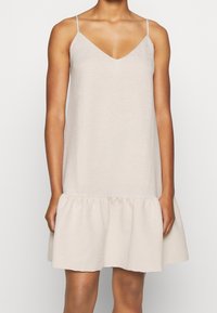 Kvinna i en ärmlös ljusbeige klänning med tunna band och volangkant står framför en enfärgad vit bakgrund.