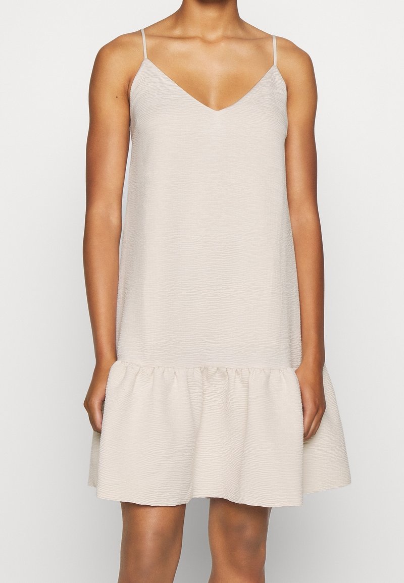 Kvinna i en ärmlös ljusbeige klänning med tunna band och volangkant står framför en enfärgad vit bakgrund.