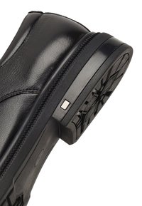 Zwarte leren enkellaars met een gladde afwerking, rubberen zool met een gebogen grippatroon en een klein metalen detail aan de zijkant.