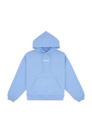Femme portant un sweat à capuche bleu clair, une mini-jupe asymétrique grise et des chaussures à talons transparents, debout devant un fond gris uni.