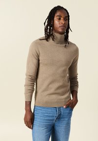 Suéter beis de cuello alto con puños y dobladillo acanalados, hecho de un material suave. Combinado con jeans de mezclilla azul claro, utilizado de manera casual.