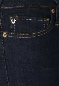 Dunkle Denim-Jeans mit gelben Nähten, einem kleinen Logo auf der Vorderseite der Tasche und einem klassischen Fünf-Taschen-Design. Glatte Textur mit schmalem Schnitt.