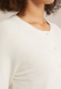 Gaišs krēms kardigāns, izgatavots no mīksta adījuma auduma, ar apaļu izgriezumu un diviem priekšējiem pogām. Redzama ribota tekstūra gar apmalēm.