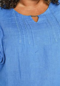Personne portant un chemisier bleu avec des détails en dentelle verticale et une petite agrafe métallique au niveau de l'encolure, montrant le haut de la poitrine et les épaules.